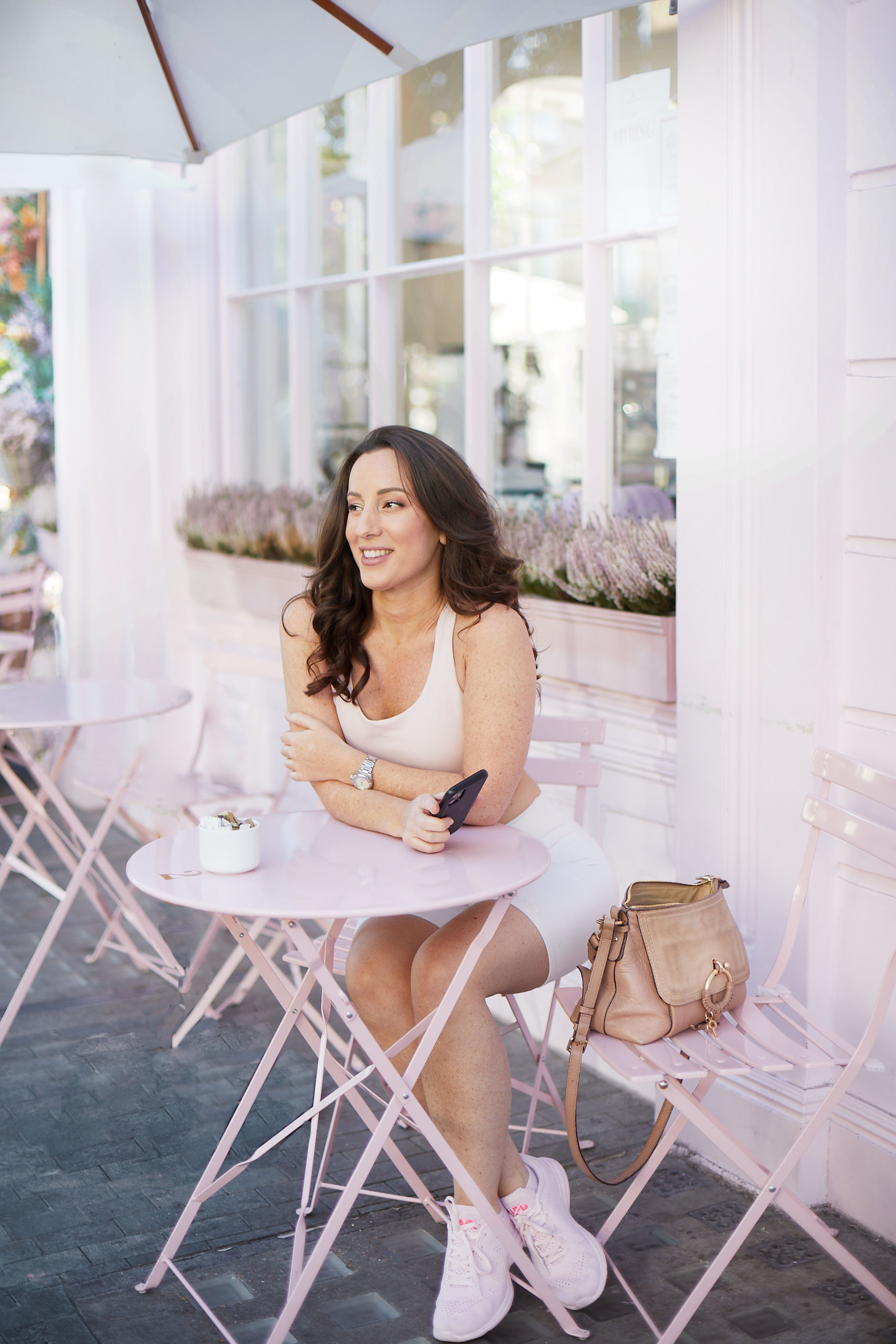 Brunette wearing fitness clothes sitting in pink cafe holding phone